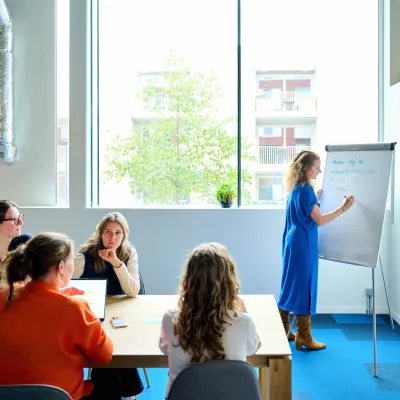 De copywriters van Bliksem Schrijfbureau zitten in meeting aan tafel, terwijl Marlies rechtstaand notities neemt op een chart board